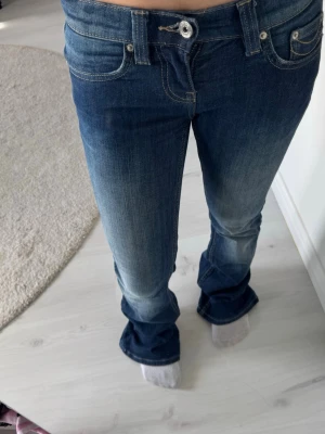 Blå bootcut jeans med slitningar - Säljer ett par blå bootcut jeans med snygga slitningar och ljusare partier framtill. Jeansen har låg midja, klassiska fem fickor och stängs med knapp och dragkedja. Perfekta för en avslappnad och trendig look.