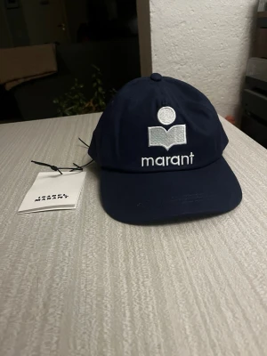 Mörkblå keps från Isabel Marant - Snygg mörkblå keps från Isabel Marant med broderad logotyp och text framtill. Klassisk böjd skärm och justerbar rem bak. Tillverkad i bomull för en bekväm och stilren look. Perfekt accessoar för att lyfta din outfit.