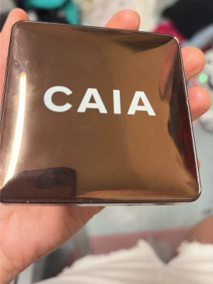 Bronzer från CAIA i guldig dosa - Snygg bronzer från CAIA i en fyrkantig, spegelblank guldig dosa med vit logga på locket. Perfekt för att ge ansiktet en solkysst look och highlighta kindbenen. Praktisk att ha med i väskan och enkel att applicera.
