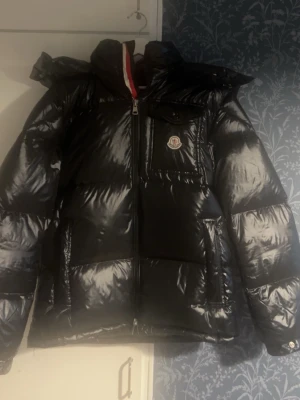 Svart glansig dunjacka från Moncler - Säljer en svart, glansig dunjacka från Moncler med huva och dragkedja framtill. Jackan har en bröstficka med Moncler-logga, stora fickor och klassisk serietecknad tvättlapp på insidan. Perfekt för kalla vinterdagar och riktigt snygg streetstil.
