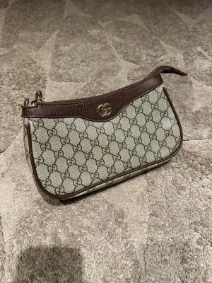 Gucci beige GG canvas handväska - Snygg beige handväska från Gucci i klassiskt GG canvasmönster med bruna läderdetaljer och guldfärgad GG-logga. Väskan har en mjuk, avlång form och dragkedja upptill. Perfekt för dig som gillar lyxiga accessoarer med ikonisk design. OBS kommer utan band