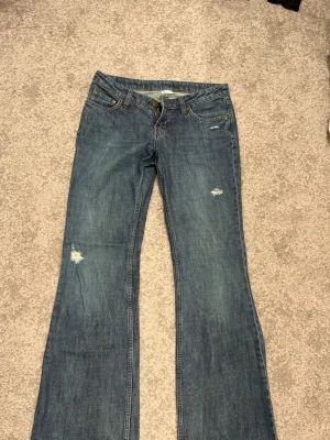 Blå bootcut jeans med slitningar - Säljer ett par blå bootcut jeans med snygga slitningar på låret och vid fickan. Jeansen har klassisk femficksmodell och låg midja. Perfekta för en avslappnad och trendig look.