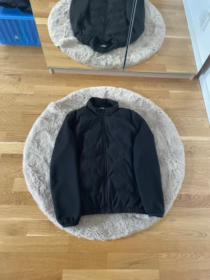 Zara Svart vindjacka med dragkedja - Zara Stilren svart vindjacka med hel dragkedja framtill och elastiska muddar vid ärmslut och midja. Jackan har en enkel design utan synliga loggor och är tillverkad i ett lätt, vindtätt material. Perfekt för blåsiga dagar och enkel att matcha med olika outfits.