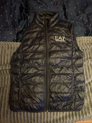 Svart dunväst från Emporio Armani - Snygg svart dunväst från Emporio Armani EA7 med quiltad design och hög krage. Västen har dragkedja framtill och logga på bröstet.