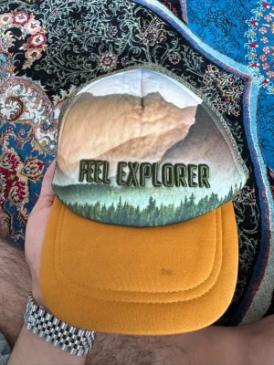 Feel Explorer truckerkeps med naturmotiv - Säljer en unik truckerkeps med broderad text 'FEEL EXPLORER' och naturmotiv framtill. Skärmen är senapsgul och baksidan är i grön mesh med justerbart spänne. Perfekt för dig som gillar outdoor-vibbar och streetstyle.