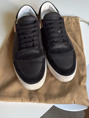 Burberry svarta sneakers med rutigt mönster - Snygga svarta sneakers från Burberry med klassiskt rutigt mönster baktill. Skorna har ovandel i mocka och textil, vita sulor och svarta skosnören. Perfekta för dig som vill ha en stilren och exklusiv look . Köpta på vinted för 3500, säljer de nu för att de blivit för små. Tvättas vid eventuellt köp!😄