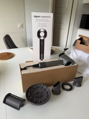 Dyson Supersonic hårfön med tillbehör - Säljer en Dyson Supersonic hårfön i grå metallic med flera olika munstycken och diffuser. Hårfönen har en modern, rund design och är tillverkad i plast och metall. Perfekt för snabb och skonsam hårtorkning med avancerad teknik.