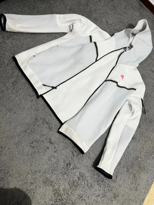 Vit softshell vindjacka med huva - Säljer en vit softshell vindjacka från Peak Performance med svarta detaljer och dragkedja. Jackan har huva, långa ärmar och en liten rosa logga på bröstet. Materialet är mjukt och stretchigt, perfekt för blåsiga dagar. Snygg passform och sportig look.