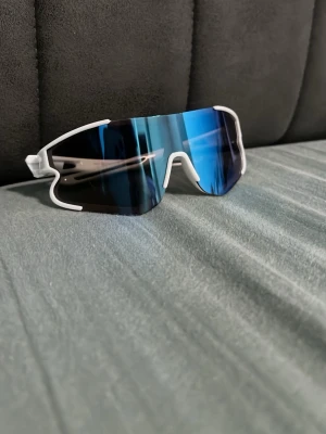 Vita sportiga solglasögon med blå spegelglas - Säljer ett par sportiga solglasögon med vit båge och stora blå spegelglas. Glasögonen har en futuristisk, svepande form och passar perfekt för en aktiv livsstil. Snygg accessoar för dig som gillar att sticka ut med din stil. Kostar 2000kr men säljs för 430kr prisförslag 