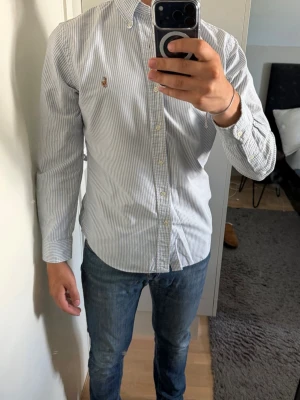 Randig skjorta från Ralph Lauren - Klassisk vit skjorta med tunna blå ränder från Ralph Lauren. Skjortan har lång ärm, knappar framtill och en broderad logga på bröstet. Tyvärr små slitningar vid kragen men förutom det är den helt perfekt. Tillverkad i bomull och har en normal passform. Perfekt för en stilren look.