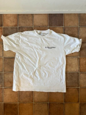 Vit Le Chaux-de-Fonds t-shirt - Vit t-shirt från Le Chaux-de-Fonds med tryck av fem klockor på ryggen och liten logga på bröstet. Klassisk passform och rund halsringning. Perfekt för dig som gillar unika prints och schweizisk stil.