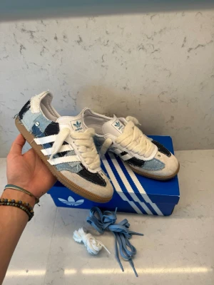 Adidas Samba kojima  - Unika Adidas Samba kojima i patchwork denim med blå, svart och vita toner. Skorna har klassiska vita stripes, beige mockadetaljer och gummisula. Levereras med extra skosnören i blått och vitt. Perfekt för dig som vill sticka ut med en retroinspirerad look.