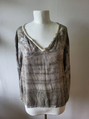  Blus med v-ringning och mönster st M - Luftig beige blus med v-ringad hals och subtilt blommigt mönster i silver. Långa ärmar och "oljebehandlade" ytor. avslappnad och trendig look. 