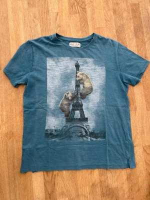 Blå t-shirt med björnar från Zara - Blå t-shirt från Zara Boys med coolt tryck av två björnar som klättrar på Eiffeltornet. Klassisk rund hals och korta ärmar. Tillverkad i mjuk bomull som känns skön mot huden. Perfekt för dig som gillar unika motiv och vill sticka ut lite extra. lite sönder längst ner se bild 