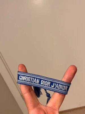 Blått armband Christian Dior J'ADIOR - Snyggt blått tygarmband från Christian Dior med texten 'J'ADIOR' broderad i vitt. Armbandet har justerbara knytband med tofsar i ändarna och en cool, ungdomlig design. Perfekt accessoar för att lyfta din outfit.