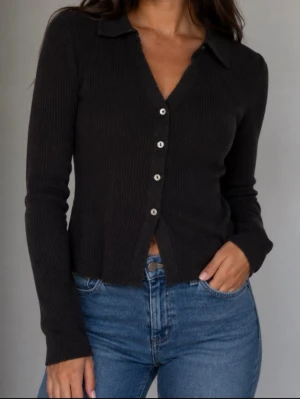 Svart ribbad långärmad topp - Stilren svart ribbad topp med lång ärm och knappar framtill. Djup v-ringning och klassisk krage ger en snygg och enkel look. Perfekt att matcha med jeans eller kjol för en trendig outfit. Pristagare saknas men helt ny