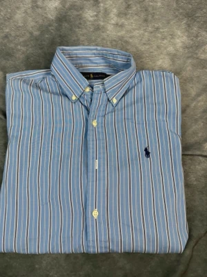 Blårandig skjorta från Ralph Lauren - Klassisk blårandig skjorta från Ralph Lauren med button-down krage och lång ärm. Skjortan har vita och ljusblå ränder samt den ikoniska mörkblå Polo-loggan broderad på bröstet. Tillverkad i bomull för en skön och luftig känsla.