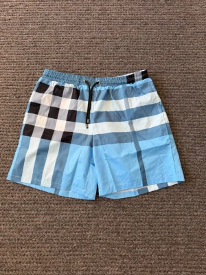 Burrberry badbyxor - Snygga badbyxor i ljusblått med klassiskt rutmönster i svart, vitt och mörkblått. De har elastisk midja med svart dragsko och metalländar. Materialet är snabbtorkande polyester, perfekt för stranden eller poolen.