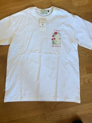 Vit Casa Blanca t-shirt med rosor - HELT NY Vit t-shirt från Casa Blanca med tryck av röda rosor och landskap både fram och bak. T-shirten har rund hals, korta ärmar och en relaxed passform. Perfekt för dig som gillar stilrena plagg med unika detaljer.