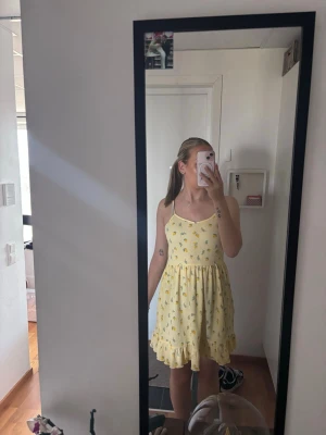 Gul blommig sommarklänning - Söt gul kortklänning med smala axelband och volangkant nertill. Klänningen har ett blommigt mönster i vitt och grönt. Aldrig använd. 