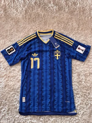 Blå Sverige matchtröja Adidas - Säljer en blå Sverige matchtröja från Adidas med gula detaljer, nummer 17 och klassiskt mönster. Tröjan har korta ärmar, V-ringning och gula ränder på axlarna. Broderad landslagssköld och FIFA-märke på ärmen. Perfekt för fotbollsfans!
