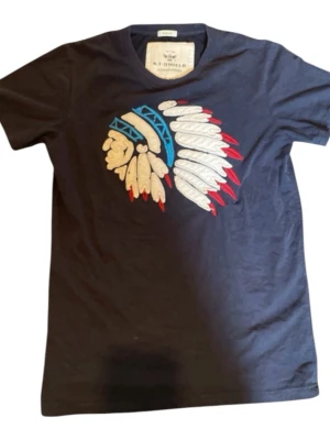 Svart t-shirt KT Shield med tryck - Mörkblå t-shirt från KT Shield med färgglatt tryck av ett huvudbonadsmotiv i vitt, blått, rött och beige på bröstet. Klassisk rund hals och korta ärmar. Tillverkad i mjuk bomull för en bekväm känsla. Storlek M, och ör vintage 