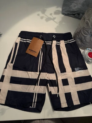 Burberry badshorts med rutigt mönster - Snygga badshorts från Burberry i marinblått och beige med klassiskt rutigt mönster. Shortsen har resår i midjan och dragsko för bästa passform. Tillverkade i snabbtorkande material, perfekta för stranden eller poolen.
