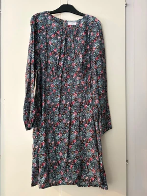 Blommig långärmad klänning - Supersöt blommig klänning med lång ärm och rund halsringning. Mönstret går i grönt, rosa, rött och vitt och ger en fräsch vibe. Klänningen har en avslappnad passform och är perfekt för vår och sommar. Tillverkad i mjukt tyg som känns skönt mot huden. Storlek 48. Från Holly & Whyte, Lindex.