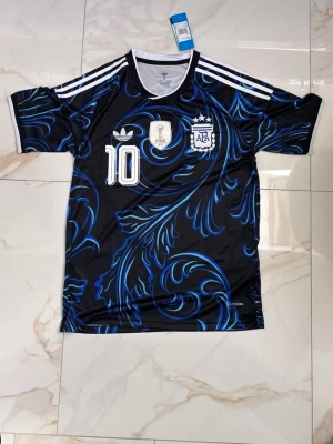 Argentina Messi Adidas matchtröja - Snygg svart och blå Argentina matchtröja från Adidas med Messi och nummer 10 på ryggen. Tröjan har vita detaljer, FIFA-märke och AFA-logga på bröstet samt ett unikt blått mönster över hela plagget. Perfekt för fotbollsfans som vill sticka ut på planen.