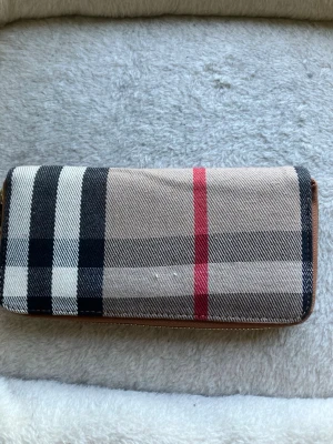 Burberry plånbok med rutigt mönster - …Går att pruta såklart…Säljer en klassisk Burberry plånbok med det ikoniska beige, svarta och röda rutmönstret. Plånboken har dragkedja i guld och detaljer i brunt läder. Perfekt för dig som vill ha en stilren och tidlös accessoar.