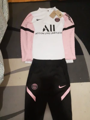 PSG träningsset Nike rosa/svart - Snyggt PSG träningsset från Nike med långärmad vit tröja med rosa och svarta detaljer samt svarta byxor med rosa inslag. Tröjan har Paris Saint-Germain-logga och sponsortryck, byxorna har PSG-märke och Nike-logga. Perfekt för fotbollsträning eller chill.