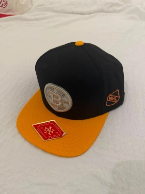Vintage American Needle svart, gul keps - Svart keps med en gul skärm. Framsidan pryds av en broderad logotyp i silver och svart. På sidan finns en orange NHL-logotyp. Kepsen har en justerbar rem baktill för passform.