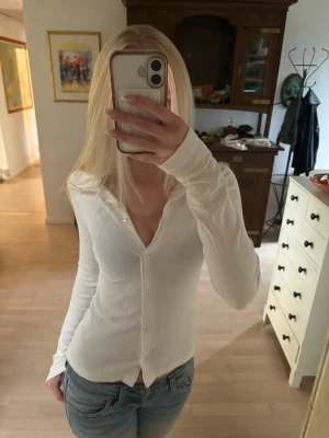 Vit ribbad långärmad tröja  - Säljer en vit ribbad tröja med v-ringning och knappar framtill. Tröjan har lång ärm och en figurnära passform som ger en clean och stilren look. Perfekt att matcha med jeans eller kjol för en enkel men snygg outfit. Tröjan är skjort liknande med stretchigt tyg. 