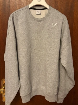 Grå oversized sweatshirt från Gymshark - En stilren grå sweatshirt från Gymshark med rund halsringning och diskret logga på bröstet. Tröjan har långa ärmar, ribbade muddar och är gjord i mjukt bomullsmaterial. Perfekt för en avslappnad och sportig look.