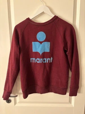 Vinröd sweatshirt från Isabel Marant - Säljer en vinröd sweatshirt från Isabel Marant med stort blått logotryck på ryggen. Tröjan har rund halsringning, raglanärmar och ribbade muddar. Perfekt för dig som gillar stilrena och trendiga plagg. Materialet är mjukt och bekvämt.