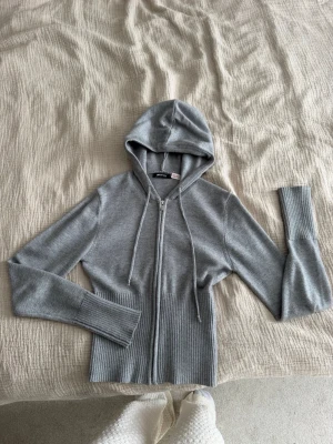 Grå zip hoodie från Gina Tricot - Säljer en grå hoodie från Gina Tricot med dragkedja framtill och huva med dragsnören. Tröjan har ribbade muddar och midja, samt långa ärmar. Perfekt för en chill och stilren look. Materialet är mjukt och stretchigt, vilket gör den superbekväm.
