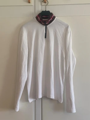 Hugo Boss Vit långärmad tröja med dragkedja - Hugo boss vit långärmad tröja med hög krage och dragkedja framtill. Kragen har röda och svarta detaljer samt textmönster. Tröjan är rak i modellen och tillverkad i ett mjukt material, perfekt för en avslappnad stil.