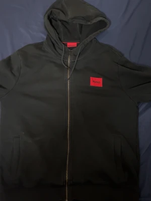 Svart hoodie med dragkedja från Hugo - Svart hoodie från Hugo med dragkedja framtill och röd logotyp-patch på bröstet. Klassisk huva med dragsko och två fickor på sidorna. Perfekt för en chill och stilren look. Materialet känns mjukt och bekvämt. Storlek L men passar M