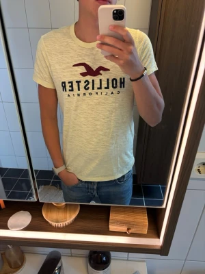 Gul Hollister t-shirt i bomull - Fräsch gul t-shirt från Hollister med klassisk logga och text i vinrött framtill. Skön passform och tillverkad i mjuk bomull. Perfekt till jeans eller shorts för en avslappnad stil.