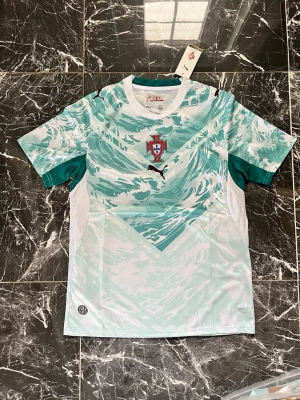Portugal Puma matchtröja turkos - Snygg Portugal matchtröja från Puma i turkos och vitt med vågigt mönster. Tröjan har rund hals, korta ärmar och Portugal-emblemet på bröstet. Tillverkad i lätt och ventilerande funktionsmaterial, perfekt för fotboll eller träning.