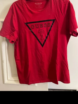 Röd t-shirt från Guess med logga - Snygg röd t-shirt från Guess med den klassiska triangelloggan tryckt i svart på bröstet. T-shirten har rund halsringning och korta ärmar. Tillverkad i mjuk bomull som känns skön mot huden. Perfekt för en trendig och avslappnad stil.