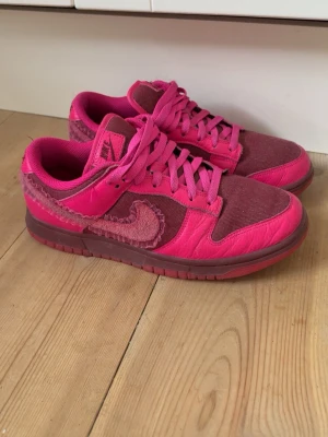 Nike Dunk Low ”Valentine’s day” - Unika Nike Dunk Low sneakers med vinröd och rosa med mockadetaljer och textil på ovandelen. Skorna har klassisk låg siluett, rosa snörning och en cool, ruffad Swoosh på sidan. Perfekta för dig som vill sticka ut med färg och stil.