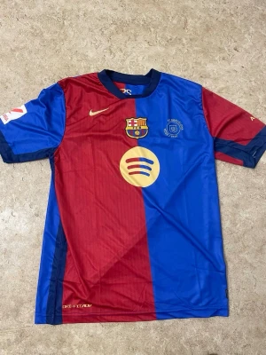 FC Barcelona matchtröja Nike röd/blå - Säljer en officiell FC Barcelona matchtröja från Nike i rött och blått med klubbmärke, Spotify-logga och UNHCR-tryck. Tröjan har Lamine Yamal och nummer 19 på ryggen. Tillverkad i lätt, ventilerande material med Dri-FIT ADV-teknologi och korta ärmar.