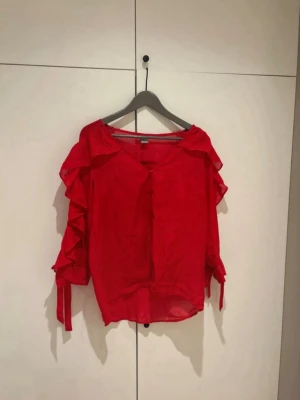 Röd blus med volangärmar - Snygg röd blus med volangdetaljer på ärmarna och knappar framtill. Blusen har en lös passform och är tillverkad i ett lätt, tunt tyg som ger en luftig känsla. Perfekt för att addera färg till din outfit.