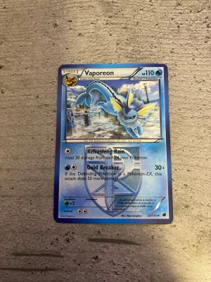 Vaporeon Pokémonkort - Säljer ett Vaporeon Pokémonkort med blå bakgrund och detaljer i silver och gult. Kortet har 110 HP och visar Vaporeon i en stadsmiljö. Det är från serien 2011 och har attacker som Refreshing Rain och Gold Breaker. Perfekt för samlare eller Pokémonfans.