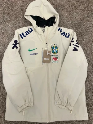 Brasil windbreaker nike - Snygg vit träningsjacka från Nike med huva och dragkedja. Jackan har Brasilien CBF-logga, gröna och blå detaljer samt sponsortryck på bröst och ärmar. Perfekt för fotbollsfans som vill ha en sportig look. Tillverkad i lätt material, passar till träning eller casual.