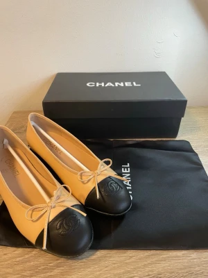 Beige och svarta ballerinaskor Chanel - Chanel ballerinaskor i beige och svart läder med klassisk rund tå och rosett framtill. Skorna har den ikoniska CC-loggan präglad på den svarta tåkappan och en låg klack. Perfekt för dig som gillar tidlös och lyxig stil.