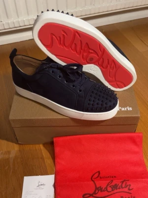 Mörkblå Louboutin sneakers med nitar - Snygga svarta sneakers från Christian Louboutin med klassisk röd sula och coola svarta nitar på tån. Skorna har en låg profil, vita sulor och är tillverkade i mocka. Perfekta för dig som vill sticka ut med exklusiv design och edgy detaljer.