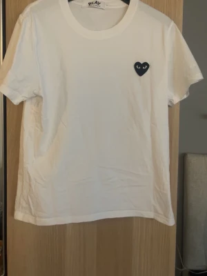 Vit t-shirt från Comme des Garçons Play - Vit t-shirt från Comme des Garçons Play med korta ärmar och klassisk rund halsringning. På bröstet finns ett svart hjärta med ögon, vilket är märkets ikoniska detalj. T-shirten är tillverkad i mjuk bomull och har en clean och stilren look.