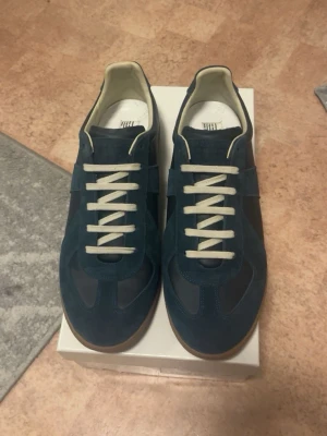 Mörkblå sneakers från Maison margiela - Snygga mörkblå sneakers med ovandel i mocka och vita skosnören. Skorna har en klassisk låg siluett och brun sula i gummi. Insidan är ljus och designen är stilren, perfekt för en trendig vardagslook.